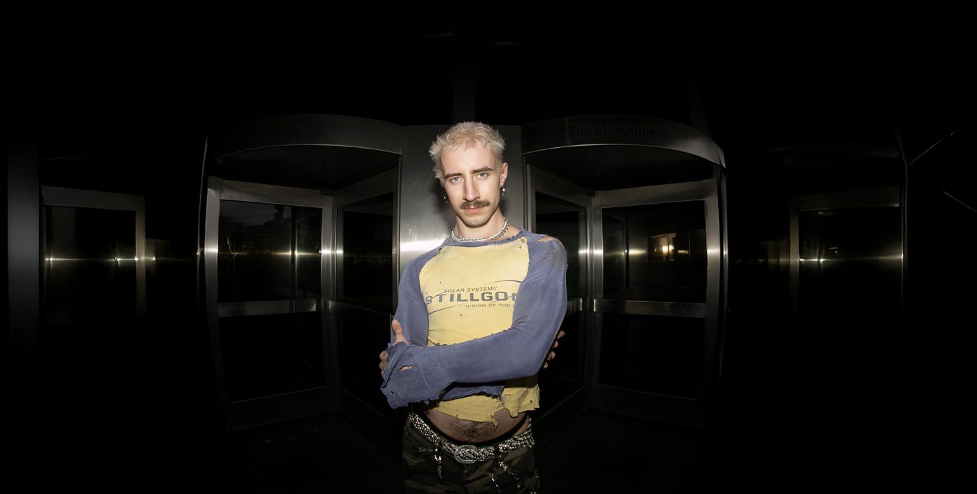 Melvin Hein in a yellow and blue shirt, arms crossed in front of a revolving door at night